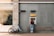 A person stands at an ATM on a city street, interacting with it. A bicycle is parked nearby against a brick building that features a large window showing indoor plants and furniture.