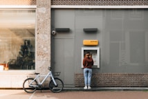 A person stands at an ATM on a city street, interacting with it. A bicycle is parked nearby against a brick building that features a large window showing indoor plants and furniture.