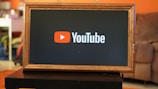A rectangular screen encased in a wooden frame displaying the YouTube logo, characterized by a red play button icon next to the word 'YouTube' on a black background. The screen is placed on a small black table in a room with an orange wall and a couch in the background.
