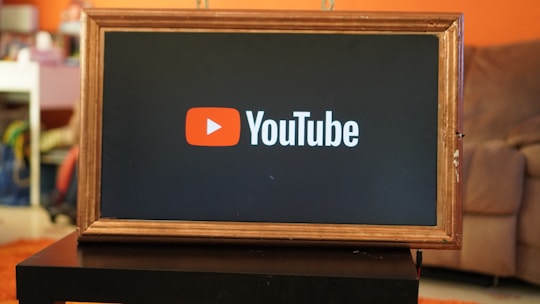 A rectangular screen encased in a wooden frame displaying the YouTube logo, characterized by a red play button icon next to the word 'YouTube' on a black background. The screen is placed on a small black table in a room with an orange wall and a couch in the background.