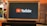 A rectangular screen encased in a wooden frame displaying the YouTube logo, characterized by a red play button icon next to the word 'YouTube' on a black background. The screen is placed on a small black table in a room with an orange wall and a couch in the background.
