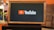 A rectangular screen encased in a wooden frame displaying the YouTube logo, characterized by a red play button icon next to the word 'YouTube' on a black background. The screen is placed on a small black table in a room with an orange wall and a couch in the background.