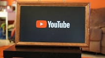A rectangular screen encased in a wooden frame displaying the YouTube logo, characterized by a red play button icon next to the word 'YouTube' on a black background. The screen is placed on a small black table in a room with an orange wall and a couch in the background.