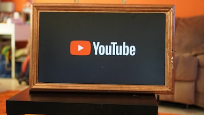 A rectangular screen encased in a wooden frame displaying the YouTube logo, characterized by a red play button icon next to the word 'YouTube' on a black background. The screen is placed on a small black table in a room with an orange wall and a couch in the background.
