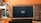 A television displaying a 'fire tv' logo is placed inside a wooden frame. The TV is positioned on a black stand in a living room with orange walls. In the background, there is a brown sofa and a white desk cluttered with various items.