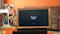 A television displaying a 'fire tv' logo is placed inside a wooden frame. The TV is positioned on a black stand in a living room with orange walls. In the background, there is a brown sofa and a white desk cluttered with various items.