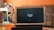 A television displaying a 'fire tv' logo is placed inside a wooden frame. The TV is positioned on a black stand in a living room with orange walls. In the background, there is a brown sofa and a white desk cluttered with various items.