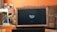 A television displaying a 'fire tv' logo is placed inside a wooden frame. The TV is positioned on a black stand in a living room with orange walls. In the background, there is a brown sofa and a white desk cluttered with various items.