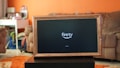 A television displaying a 'fire tv' logo is placed inside a wooden frame. The TV is positioned on a black stand in a living room with orange walls. In the background, there is a brown sofa and a white desk cluttered with various items.