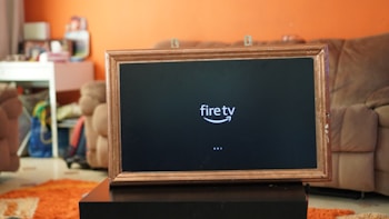 A television displaying a 'fire tv' logo is placed inside a wooden frame. The TV is positioned on a black stand in a living room with orange walls. In the background, there is a brown sofa and a white desk cluttered with various items.
