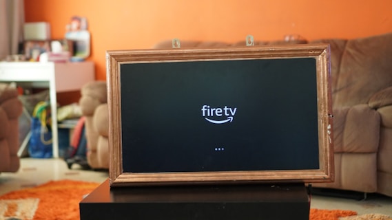 A television displaying a 'fire tv' logo is placed inside a wooden frame. The TV is positioned on a black stand in a living room with orange walls. In the background, there is a brown sofa and a white desk cluttered with various items.