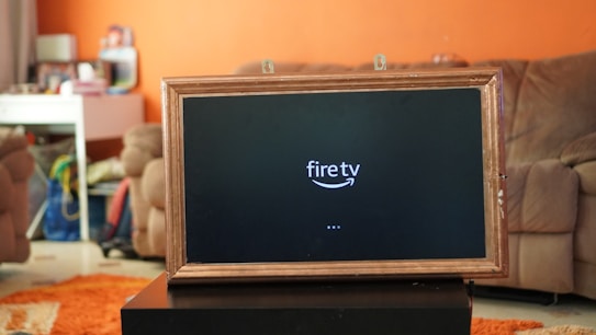 A television displaying a 'fire tv' logo is placed inside a wooden frame. The TV is positioned on a black stand in a living room with orange walls. In the background, there is a brown sofa and a white desk cluttered with various items.