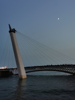 A modern bridge with a prominent angled white column supports multiple cables over calm water. The bridge is elegantly illuminated, with a crescent moon visible in the dusky sky.