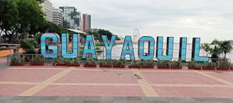 Large, colorful letters spelling 'GUAYAQUIL' are prominently displayed in a public area. The letters are set against a backdrop of tall buildings and a ferris wheel. There are plants and trees surrounding the display, adding greenery to the scene.