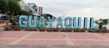 Large, colorful letters spelling 'GUAYAQUIL' are prominently displayed in a public area. The letters are set against a backdrop of tall buildings and a ferris wheel. There are plants and trees surrounding the display, adding greenery to the scene.