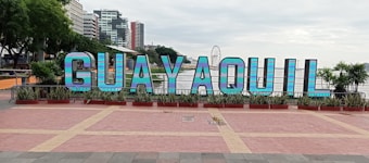 Large, colorful letters spelling 'GUAYAQUIL' are prominently displayed in a public area. The letters are set against a backdrop of tall buildings and a ferris wheel. There are plants and trees surrounding the display, adding greenery to the scene.