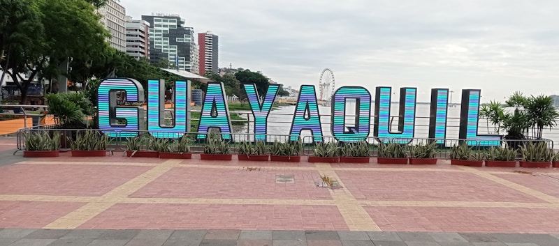 Large, colorful letters spelling 'GUAYAQUIL' are prominently displayed in a public area. The letters are set against a backdrop of tall buildings and a ferris wheel. There are plants and trees surrounding the display, adding greenery to the scene.