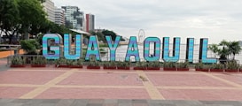 Large, colorful letters spelling 'GUAYAQUIL' are prominently displayed in a public area. The letters are set against a backdrop of tall buildings and a ferris wheel. There are plants and trees surrounding the display, adding greenery to the scene.