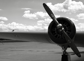 A vintage airplane engine and propeller are prominently displayed against a backdrop of a cloudy sky. The image is in black and white, emphasizing the mechanical details of the engine. The airplane wing stretches out to the left, and the concrete ground adds to the industrial feel.