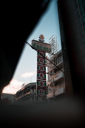 An urban scene with a tall vertical sign featuring both Thai and Chinese characters. The sign is affixed to a large building, and other buildings are visible in the background. The setting appears to be in a city as the sun is setting or rising, casting a warm glow on the environment. The sky is partly cloudy, adding to the atmospheric mood.