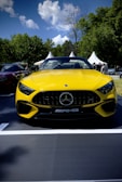 A bright yellow Mercedes-Benz AMG car is prominently displayed outdoors. The front view emphasizes its sleek design with a large grille and sharp headlights. The scene is set against a backdrop of green trees under a bright blue sky with white clouds. Several white tents and people are seen in the background.