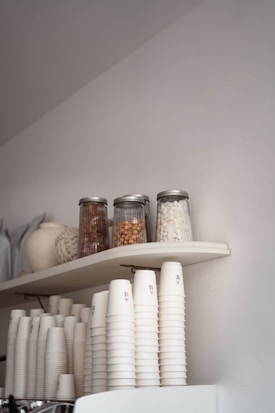 A neatly arranged set of white paper cups are stacked on a countertop beneath a shelf. The shelf holds three glass jars containing items like nuts or snacks. A larger ceramic vase and other decorative objects are partially visible beside the jars. The setting appears minimalistic and organized.