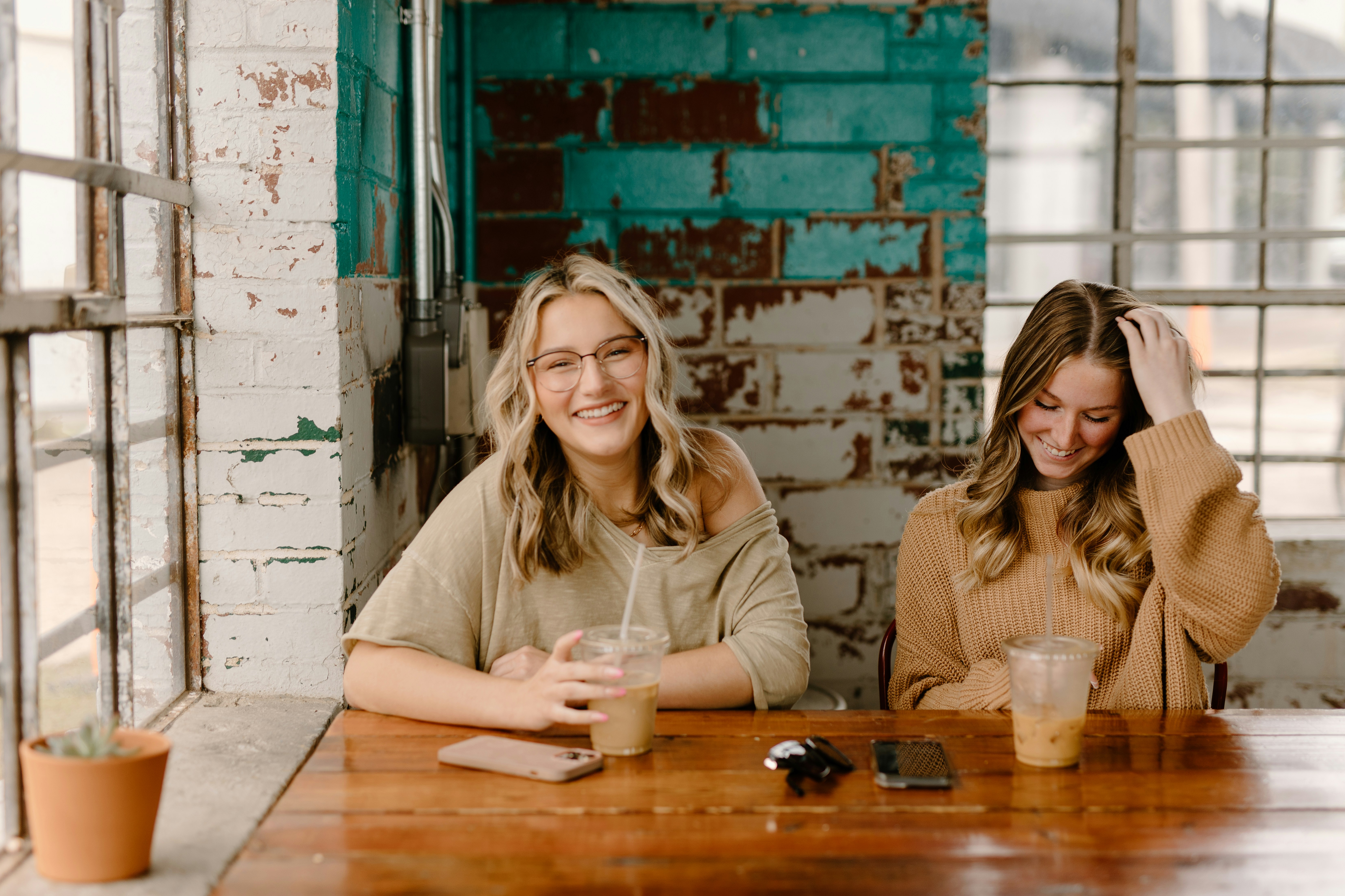 Two friends smiling and clinking coffee cups together at a café window seat
