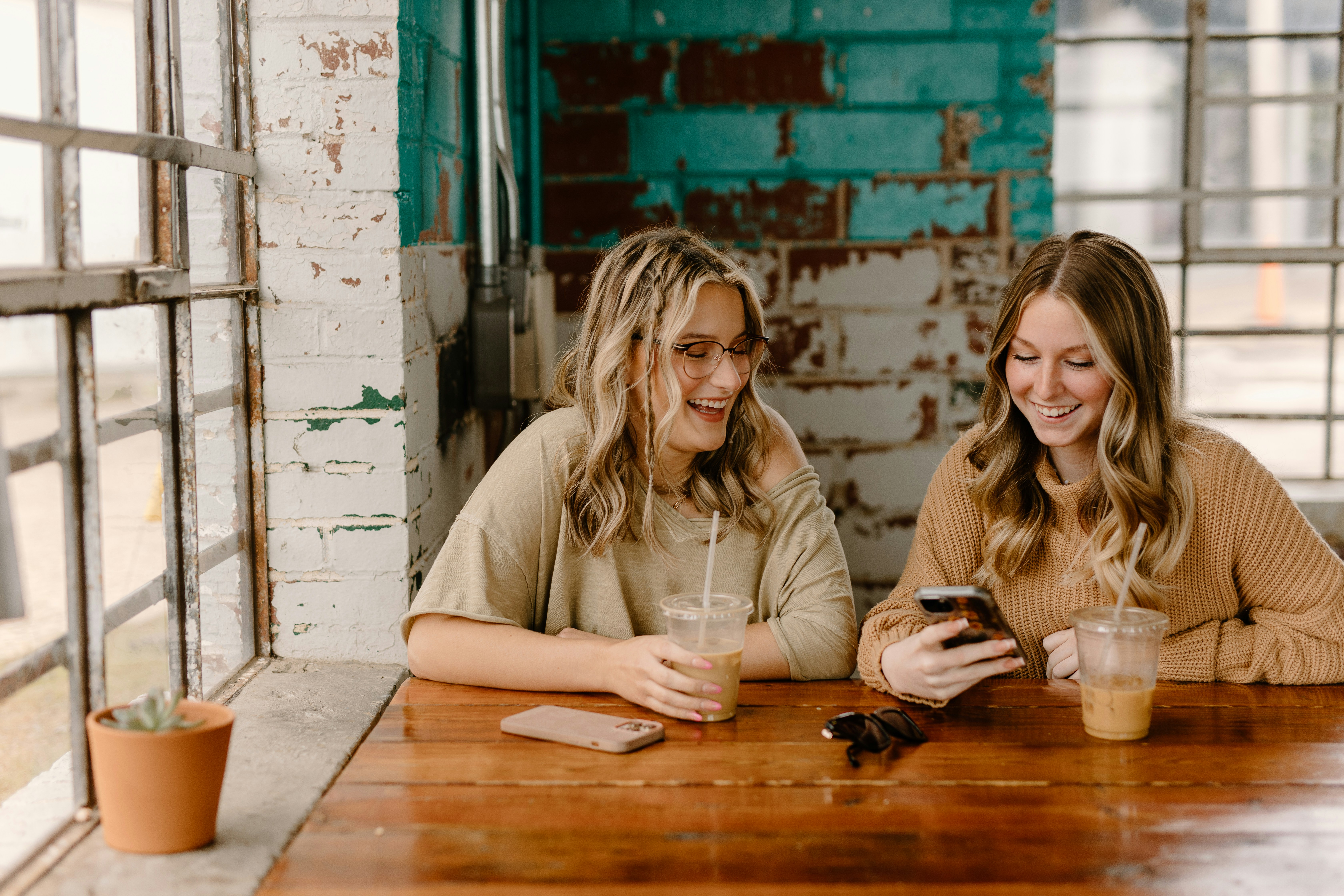 Two women with iced coffees sitting at a table laughing while looking at a phone