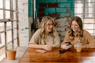 two women sitting at a table looking at a cell phone