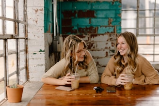 Two friends laughing over coffee at a chic café, surrounded by warm wooden tones and lush greenery.