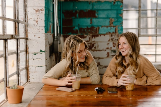 Two friends laughing together over coffee in a cozy, sunlit room filled with books and plants.