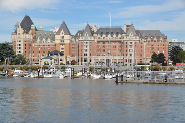 A grand, historic hotel with elaborate architecture and multiple spires is situated along a waterfront. Numerous boats are docked in the marina in front of the hotel. There are trees and greenery surrounding the building, and the sky is clear with some clouds.