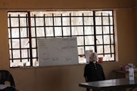 A classroom scene with a whiteboard at the front, which contains some written information and diagrams. The room has barred windows allowing natural light to come through. On the right side of the image, there is a mannequin head or a figure with a mask, positioned near the front of the classroom. Several other objects are on the desk including bottles and a container of liquid.