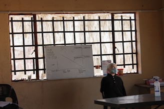 A classroom scene with a whiteboard at the front, which contains some written information and diagrams. The room has barred windows allowing natural light to come through. On the right side of the image, there is a mannequin head or a figure with a mask, positioned near the front of the classroom. Several other objects are on the desk including bottles and a container of liquid.