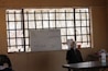 A classroom scene with a whiteboard at the front, which contains some written information and diagrams. The room has barred windows allowing natural light to come through. On the right side of the image, there is a mannequin head or a figure with a mask, positioned near the front of the classroom. Several other objects are on the desk including bottles and a container of liquid.