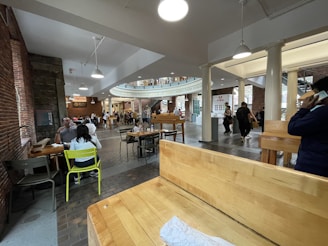 A spacious indoor area featuring a dining and gathering space with brick walls, wooden tables, and chairs. Several people are seated and walking around, with some standing in line at a vendor stall. Overhead lighting illuminates the area, and there are decorative columns and an upper floor balcony visible.