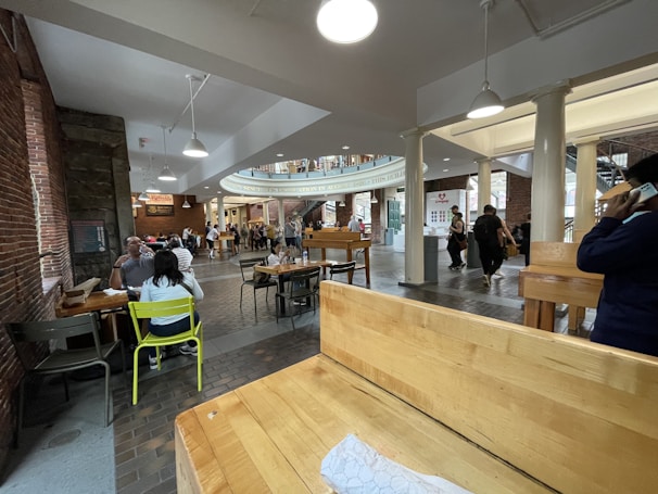 A spacious indoor area featuring a dining and gathering space with brick walls, wooden tables, and chairs. Several people are seated and walking around, with some standing in line at a vendor stall. Overhead lighting illuminates the area, and there are decorative columns and an upper floor balcony visible.