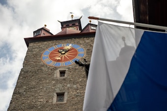 A historic clock tower with a detailed round clock face exhibits Roman numerals and a zodiac design. The tower is constructed from stone, with red-roofed structures and spires at the top. A flag in blue and white is partially visible, creating a shadow against the tower.