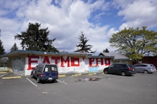 An outdoor parking area with three cars parked in front of a building featuring large red graffiti letters on a white wall. Trees with dense foliage are visible in the background, accompanied by a partly cloudy sky.