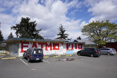 An outdoor parking area with three cars parked in front of a building featuring large red graffiti letters on a white wall. Trees with dense foliage are visible in the background, accompanied by a partly cloudy sky.