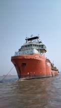 A large red ship is anchored in the water, displaying a robust structure with multiple decks and communication equipment on top. The water is calm, and another vessel is visible in the background under a clear blue sky.