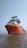 A large red ship is anchored in the water, displaying a robust structure with multiple decks and communication equipment on top. The water is calm, and another vessel is visible in the background under a clear blue sky.
