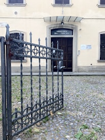 A vintage building entrance featuring a dark wooden door with a glass section labeled 'Ambulanza Med. Chirurgica.' The exterior wall is a light yellow tone and has two windows with closed wooden shutters. In front of the entrance, there is a wrought iron gate partially open, surrounded by a cobblestone path with sparse patches of grass and dry leaves.
