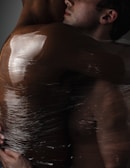 Two individuals are closely embracing, wrapped in a translucent material that partially obscures them. The lighting highlights the texture and sheen of the material, creating a sense of intimacy and vulnerability.