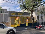 A storefront with the name 'Dog is Cool' prominently displayed in yellow letters. The entrance is secured with a mesh gate and features two bright orange banners advertising services such as 'Banho & Tosa.' A tree stands on the sidewalk next to the entrance. A white car and two red motorcycles are parked on the street in front of the building, which is surrounded by a fence with barbed wire at the top.