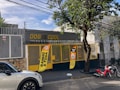 A storefront with the name 'Dog is Cool' prominently displayed in yellow letters. The entrance is secured with a mesh gate and features two bright orange banners advertising services such as 'Banho & Tosa.' A tree stands on the sidewalk next to the entrance. A white car and two red motorcycles are parked on the street in front of the building, which is surrounded by a fence with barbed wire at the top.