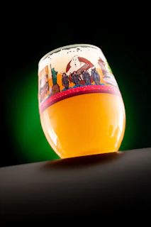A glass of beer with a frothy head, adorned with a colorful illustrated design featuring characters and a logo. The glass is tilted, set against a gradient green and black background.