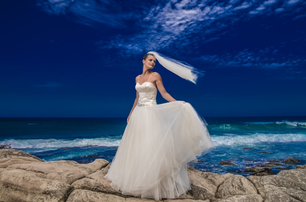 a woman in a wedding dress standing on a rock by the ocean