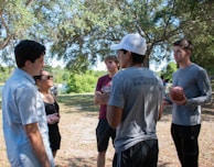 A group of young people is engaged in a conversation in a park-like setting. They stand under the shade of tall trees with sunlight filtering through the leaves. One person is holding a football, suggesting a casual or recreational gathering.