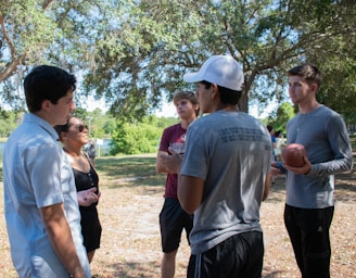 A group of young people is engaged in a conversation in a park-like setting. They stand under the shade of tall trees with sunlight filtering through the leaves. One person is holding a football, suggesting a casual or recreational gathering.